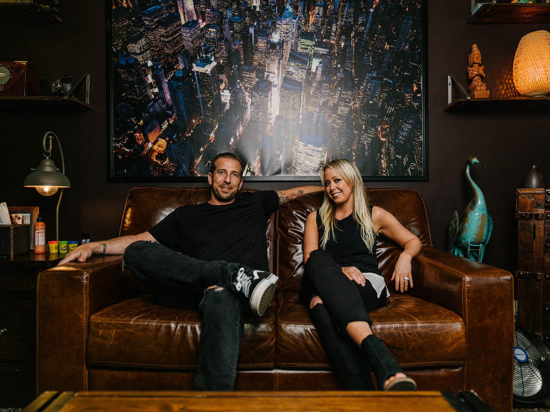 John and Michele Mopper, licensed therapists, sitting on a leather couch beneath a large NYC skyline painting, providing the best mental health approaches.